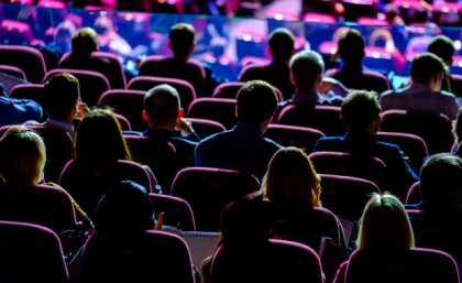 Backs of an audience in a dark conference room facing the stage.